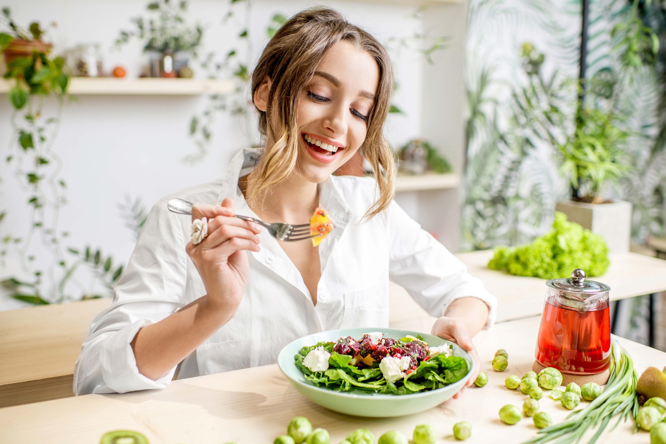 Eine Frau sitzt lachend am Tisch vor einem großen Teller Salat, im Hintergrund sind viele Pflanzen, auf dem Tisch liegt loser Rosenkohl