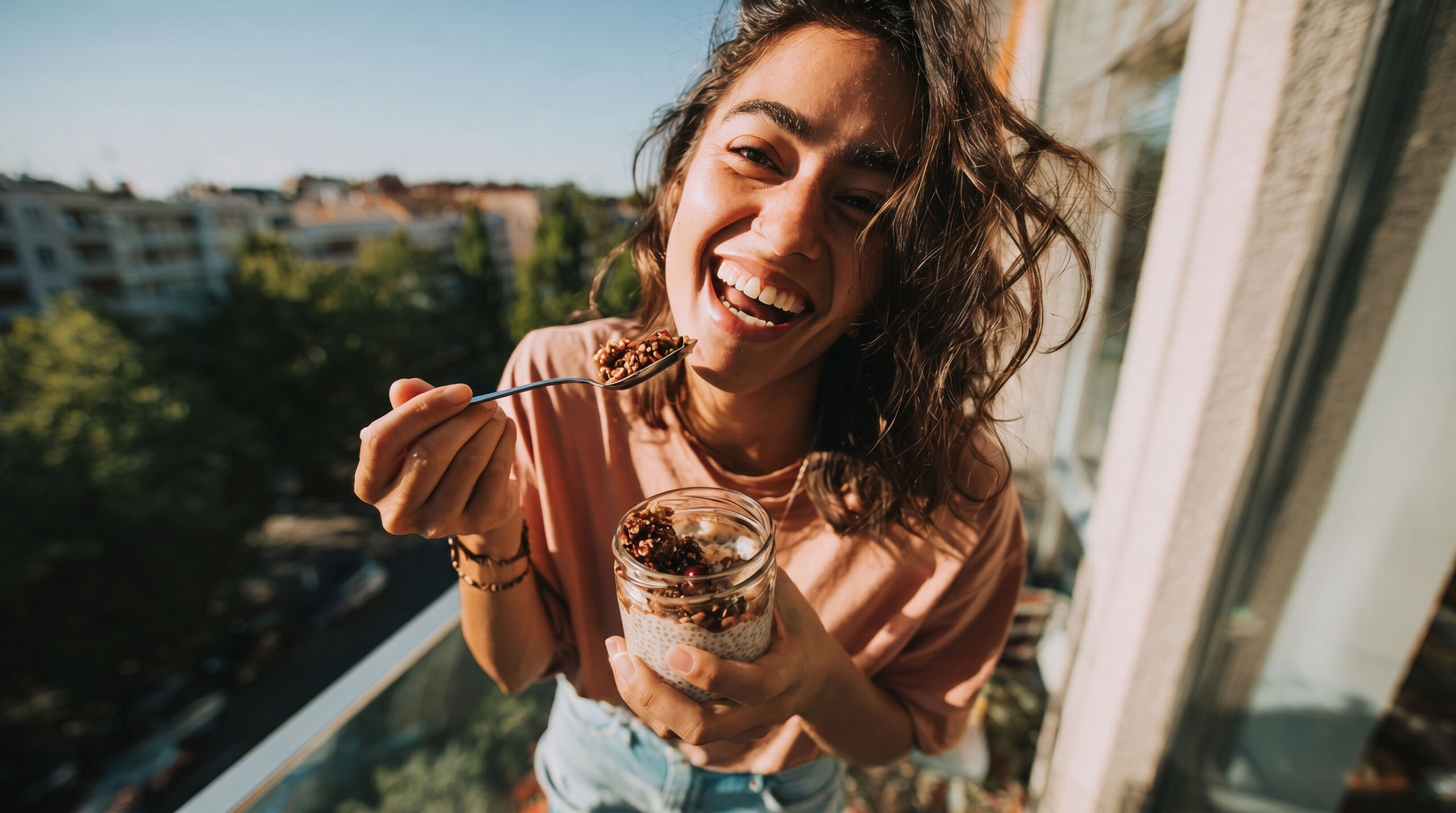 Eine Frau steht lachend auf dem Balkon und löffelt aus einem Glas mit Chiapudding und Müsli