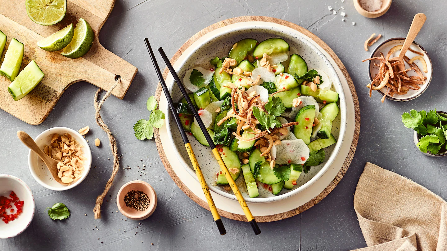 Ein gedeckter Tisch von oben fotografiert, in der Mitte ein großer Teller mit asiatischen Gurkensalat mit Erdnüssen und Chili, Essstäbchen am Tellerrand, daneben viele kleinere Gefäße mit den einzelnen Bestandteilen des Salats