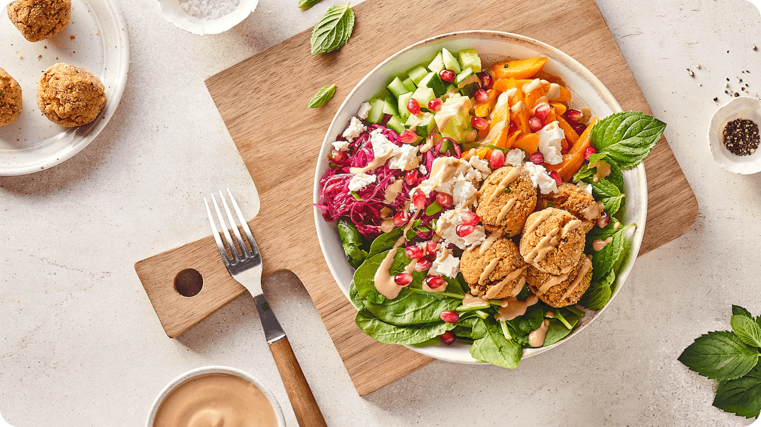 bunte falafel bowl Eine Bowl gefüllt mit bunten Zutaten wie Spinat, Rotkraut und Mango, auf einem Holzbrett stehend, daneben eine Gabel und lose Kräuter