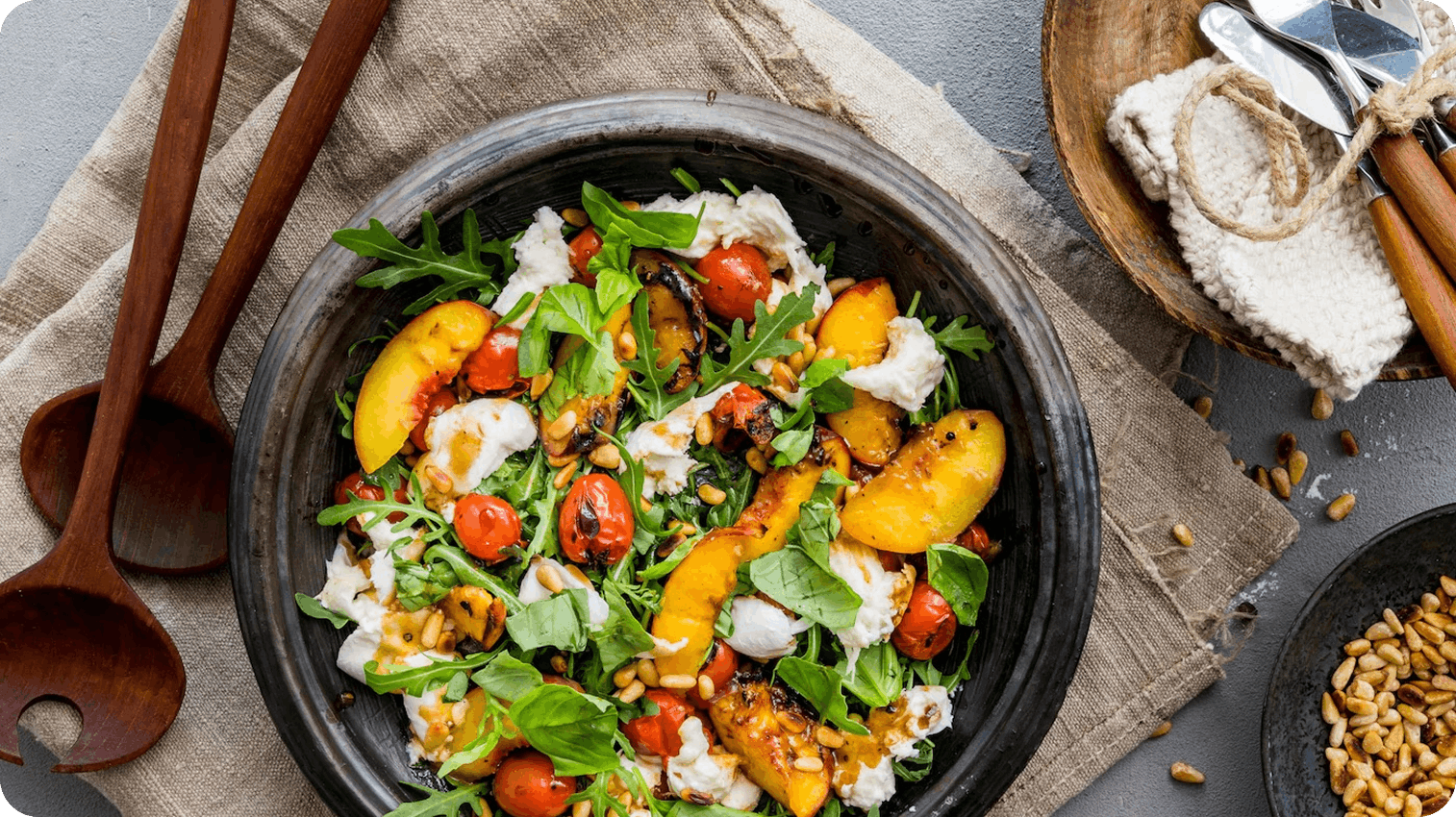 pfirsich salat Eine schwarze Schale gefüllt mit Rucola, gegrillten Pfirsichen, Tomaten und Pinienkernen, daneben drapierte Holzlöffel und Geschirrtücher