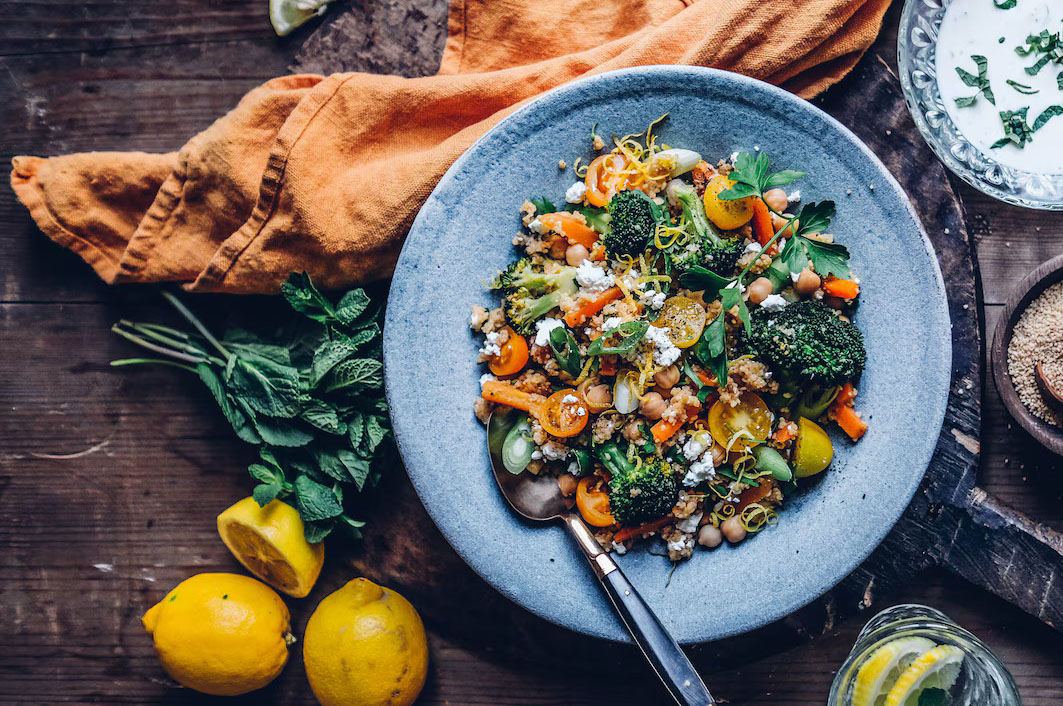 Ein Teller voll mit orangen, gelbem und grünem Gemüse auf einem Holztisch, daneben dekorativ aufgeschnittene Zitronen, etwas Minze und eine leger zusammengelegte, leuchtend orange Stoffserviette.