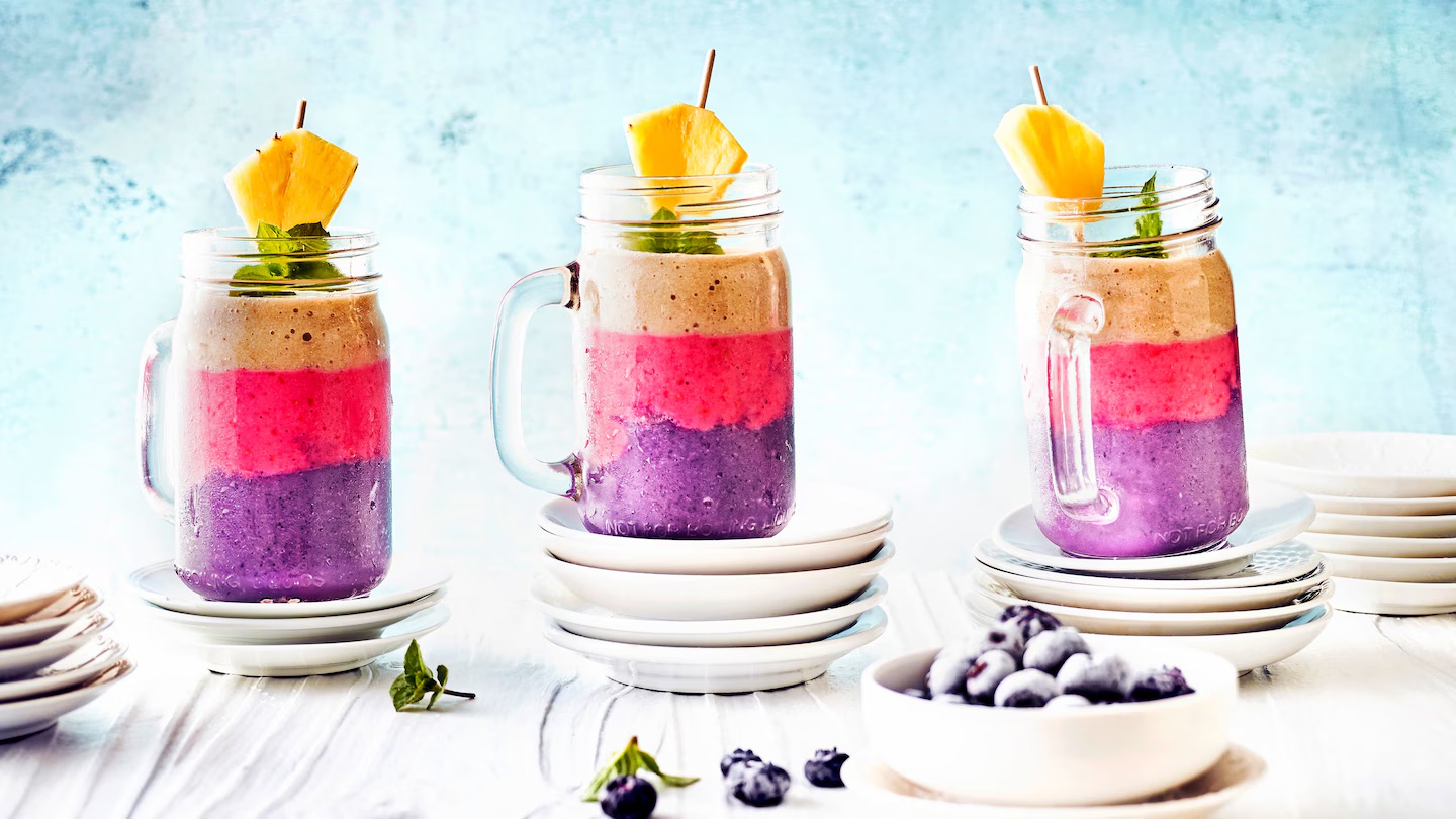 Drei Glaskrüge auf Unterteller gestapelt, darin ein Milkshake in drei Schichten mit Blaubeer-, Himbeer- und Bananengeschmack, mit einem Ananasstück und Minze dekoriert