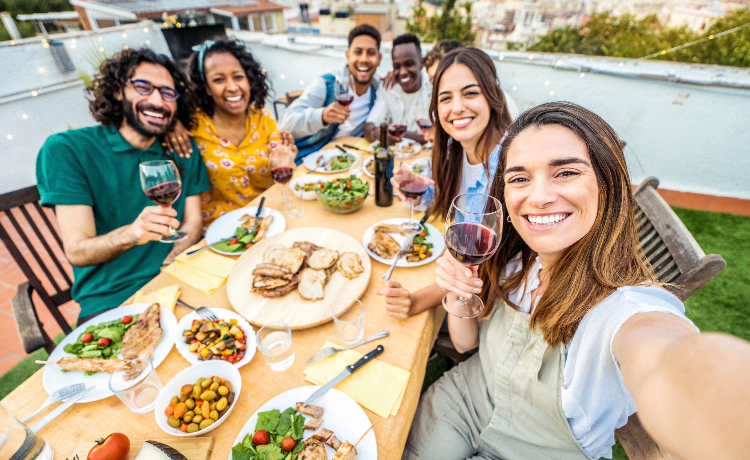Eine bunt gemischte Gruppe von Freunden sitzt gemeinsam auf einer Dachterrasse und lächelt mit Wein in die Kamera, auf dem Tisch stehen Grillfleisch und andere Speisen