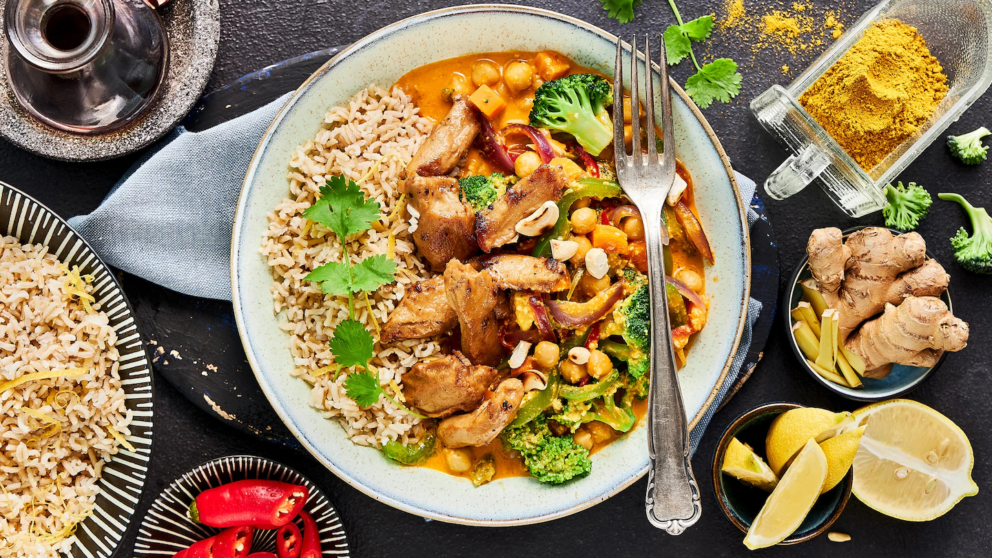 edeka-buntes-curry-mit-kichererbsen-rezept Ein Teller mit einem bunten Curry mit Vollkornreis, Hähnchen und verschiedenem Gemüse.