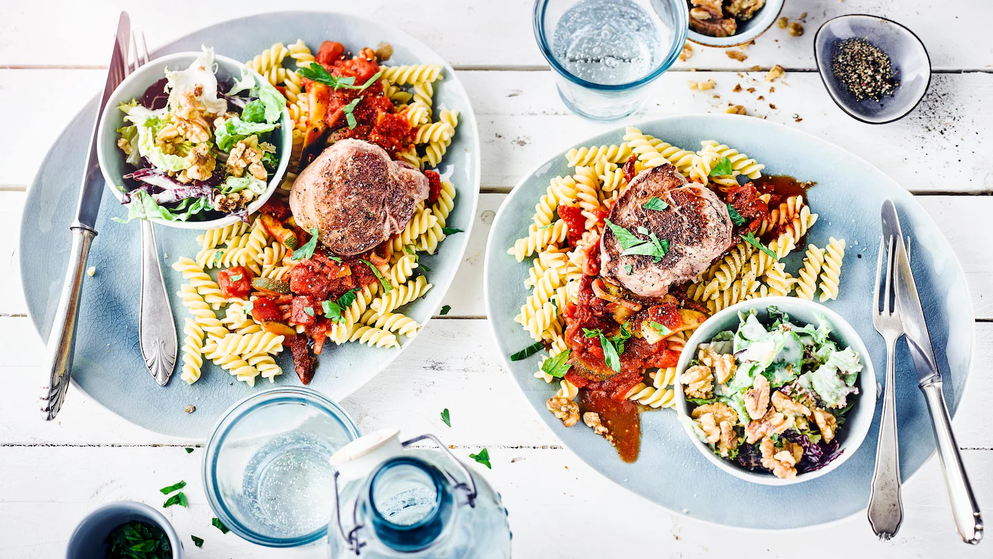 Schweinemedaillons mit Zucchini-Tomatensoße, Nudeln, Blattsalat und gerösteten Walnüssen auf einem Teller.