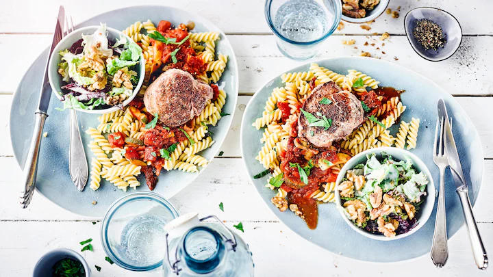 Edeka-Medaillon-Zucchini-Pfanne-Rezept Schweinemedaillons mit Zucchini-Tomatensoße, Nudeln, Blattsalat und gerösteten Walnüssen auf einem Teller.