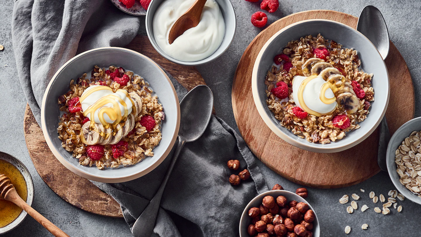 edeka-suesses-ofen-porridge-rezept Zwei Schalen mit Ofen-Porridge mit Joghurt, Himbeeren, Haselnüssen und Bananen stehen auf Holzbrettchen.