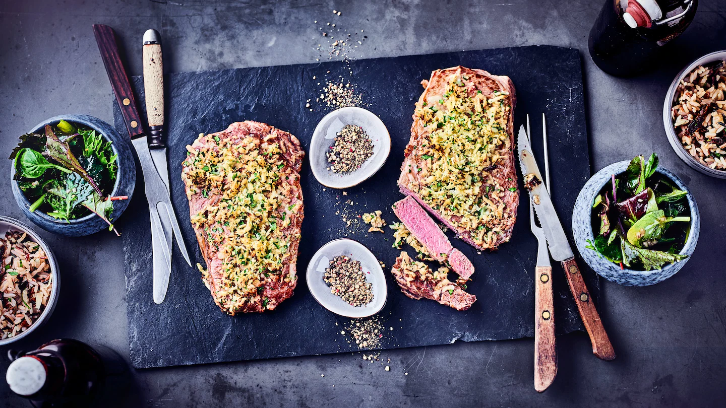 Überbackenes Rindersteak mit Käse-Kräuter-Kruste auf Schiefer, dazu Reis und Salat.