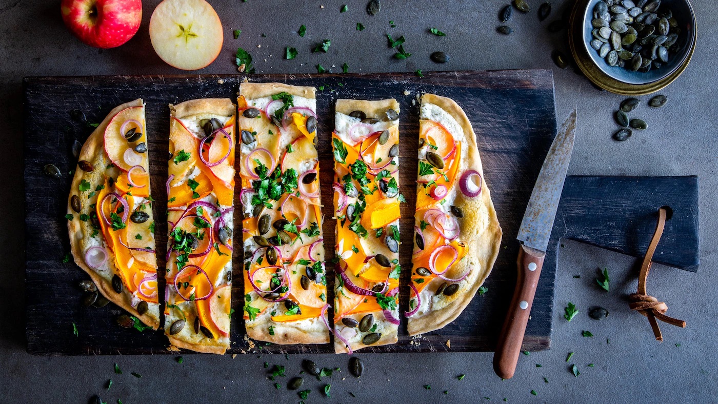 kuerbis-flammkuchen-rezept-auf-holzbrett Flammkuchen mit heller Creme, getoppt mit Kürbisscheiben, Apfel und Kürbiskernen, auf einem rustikalen Holzbrett