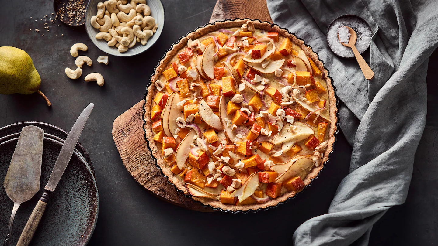 Kürbis-Birnentarte Eine Tarte mit Birnen, Kürbis und Cashewnüssen in einer Backform auf einem Holzbrett, daneben weitere Zutaten