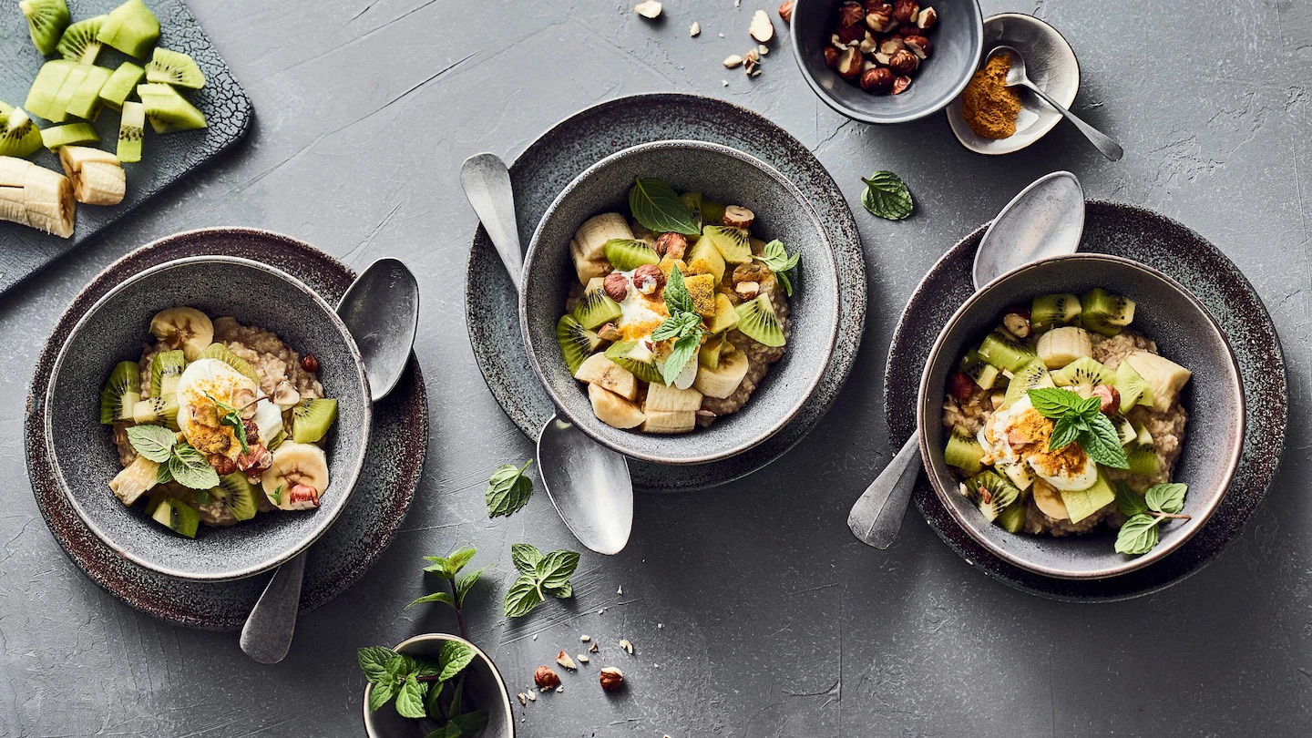 rez-edeka-kiwi-bananen-bowl-rezept-i-p Drei graue Schalen auf Untertellern gefüllt mit Poridge, Bananen, Kiwis und Nüssen. Daneben Löffel und weitere Zutaten.