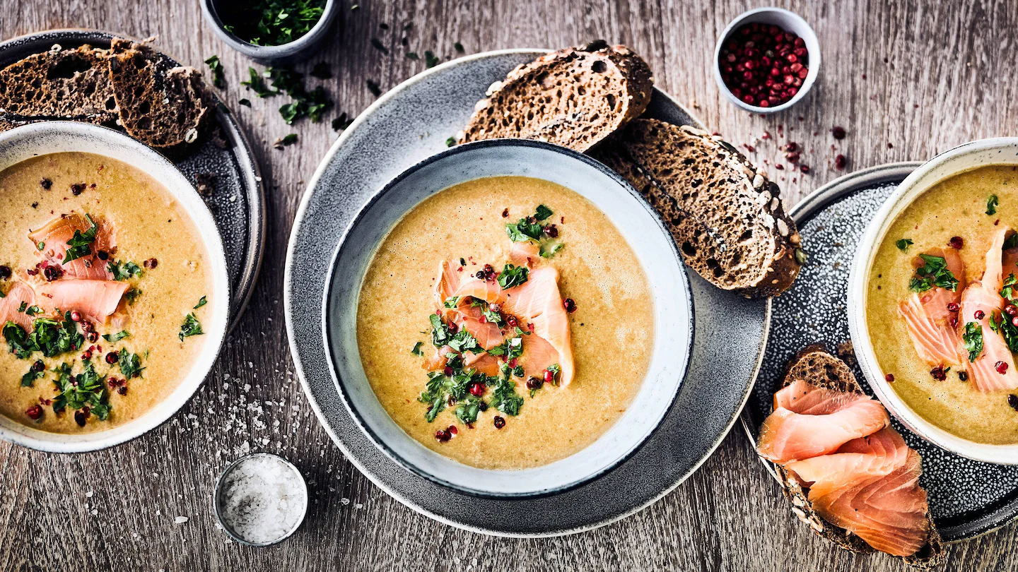 rez-edeka-pilzcremesuppe-mit-lachs-rezept-i-p-16-9 Drei Schalen mit Suppe, in der Mitte Räucherlachs und Kräuter, daneben Brot und Gewürze