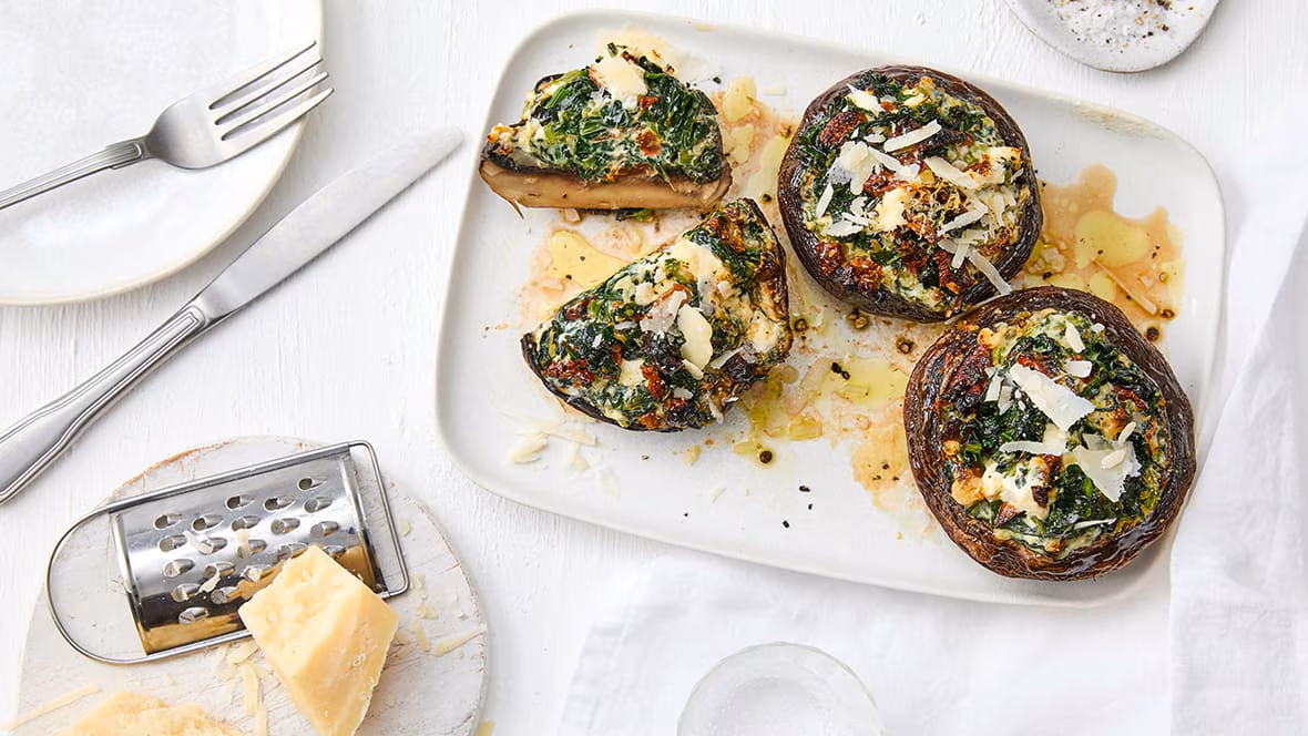 rez-edeka-portobello-pilze-mit-spinatfuellung-rezept-i-p Drei gefüllte Portobello-Pilze auf einem rechteckigen Teller, daneben Besteck und ein Parmesan mit Reibe