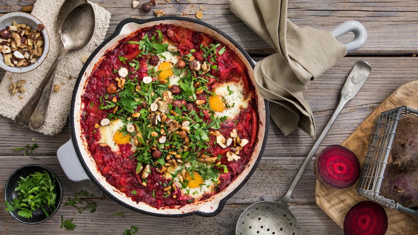 rez-edeka-rote-bete-shakshuka-rezept-q-t Eine weiße Gusseiserne Pfanne auf einem Holzuntergrund gefüllt mit rot-pinken Soße, vier Spiegeleiern, frischen Kräutern und Nüssen. Neben der Pfanne liegen verschiedene Utensilien und Zutaten.
