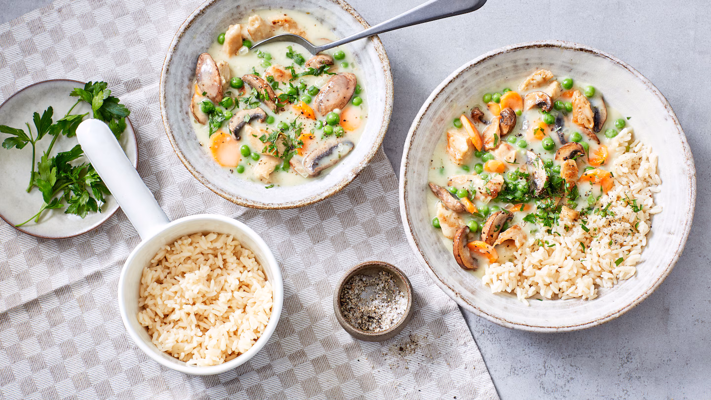 Vegetarisches Hühnerfrikassee mit Erbsen, Möhren, Champignons und Reis, angerichtet in hellem Keramikgeschirr auf einem karierten Tuch.